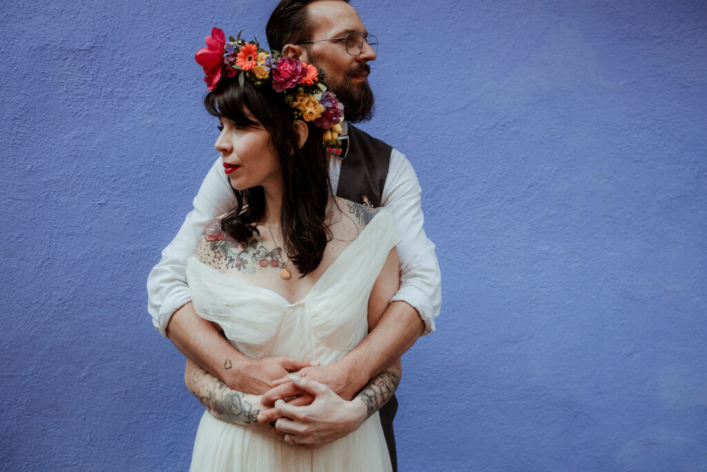 Marié dans son costume et mariée avec sa robe de mariée et sa couronne de fleurs sur la tête pour leur séance photo couple lors de leur mariage. Mur bleu et ambiance mexicaine à la Frida Kahlo