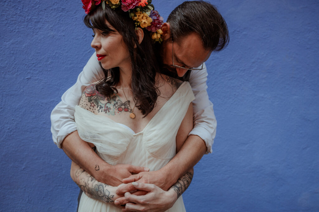 Marié dans son costume et mariée avec sa robe de mariée et sa couronne de fleurs sur la tête pour leur séance photo couple lors de leur mariage. Mur bleu et ambiance mexicaine à la Frida Kahlo